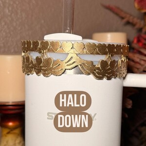 May include: A white tumbler with a gold decorative band featuring heart and leaf cutouts. The tumbler has a clear straw and the words "HALO DOWN" printed on the front. The background is a neutral color.