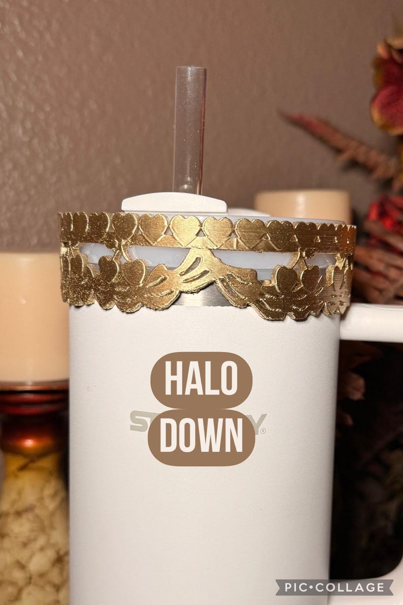 May include: A white tumbler with a gold decorative band featuring heart and leaf cutouts. The tumbler has a clear straw and the words "HALO DOWN" printed on the front. The background is a neutral color.