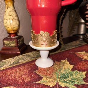 May include: A red candle in a white pedestal holder with a gold floral design. The candle holder is on a table runner with a leaf pattern. A decorative vase is in the background.