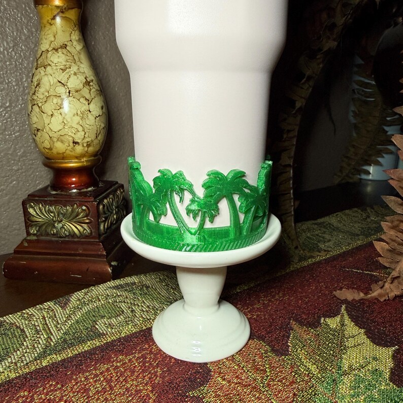 May include: A white pedestal stand featuring a decorative green wrap around the base of the bowl. The wrap displays a repeating pattern of palm trees. The stand has a smooth, solid white finish on the bowl, stem, and circular base.