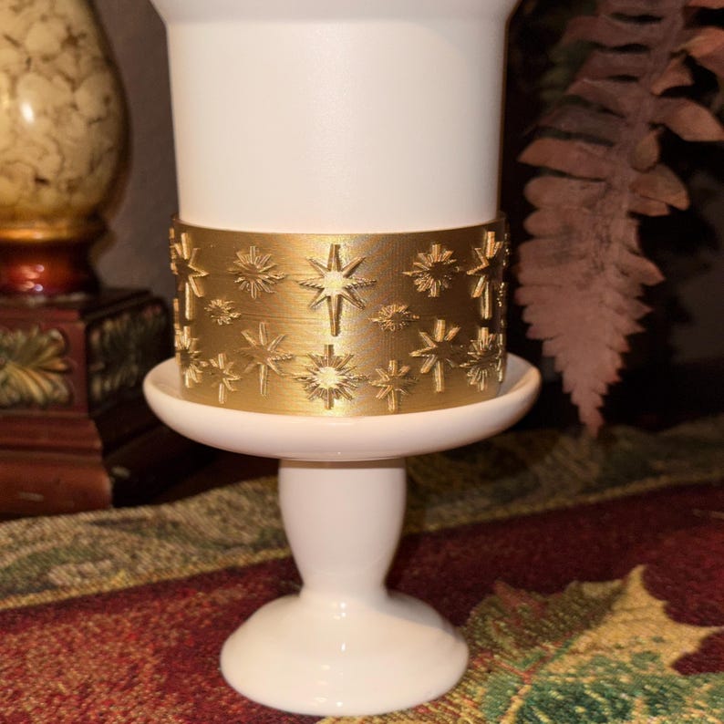 May include: A white candle holder with a gold band featuring star designs. The candle holder has a pedestal base and a wide, shallow dish to hold the candle. The gold band adds a decorative touch to the otherwise simple design.