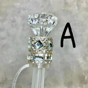 May include: A clear crystal diamond-shaped topper with a silver band decorated with rhinestones and smaller clear crystals. The topper is attached to a clear plastic rod.