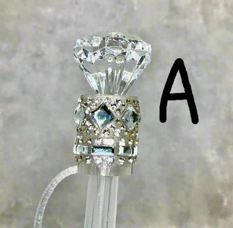May include: A clear crystal diamond-shaped topper with a silver band decorated with rhinestones and smaller clear crystals. The topper is attached to a clear plastic rod.