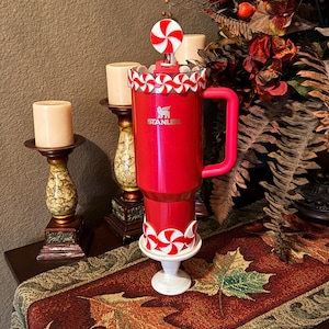 May include: A red Stanley travel mug with a candy cane theme. The mug has a red handle and a white and red candy cane design around the top and base. A candy cane topper is on the lid. The Stanley logo is visible on the front.