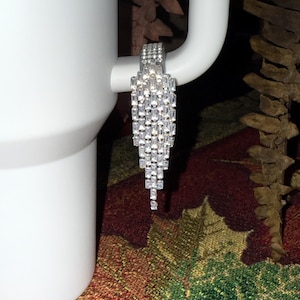 May include: A silver-toned earring with a cascade of rectangular, clear crystals. The earring is attached to a white object, possibly a mug. The crystals are arranged in a graduated, fringed design, creating a sparkling effect.