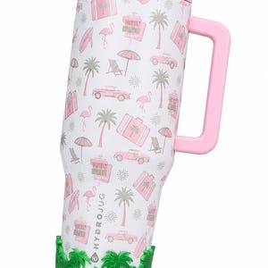 May include: A white tumbler with a pink handle and lid, featuring a repeating pattern of pink flamingos, palm trees, vintage cars, suitcases, and hotel signs. The base is encased in a green silicone sleeve with a palm tree cutout design.