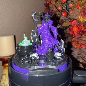 May include: A Halloween-themed decorative piece featuring a purple witch figurine with a staff, cauldron, and tombstone accents. The witch is wearing a purple dress and hat. The base is black and purple, with a spider and white roses.