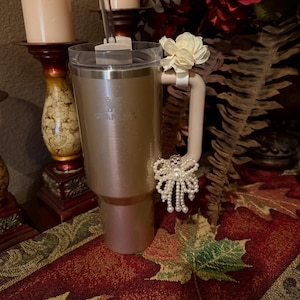 May include: A rose gold stainless steel travel mug with a white lid and a white handle. The mug has a white flower and a pearl bow attached to the handle. The mug is sitting on a table with a red and white patterned tablecloth.