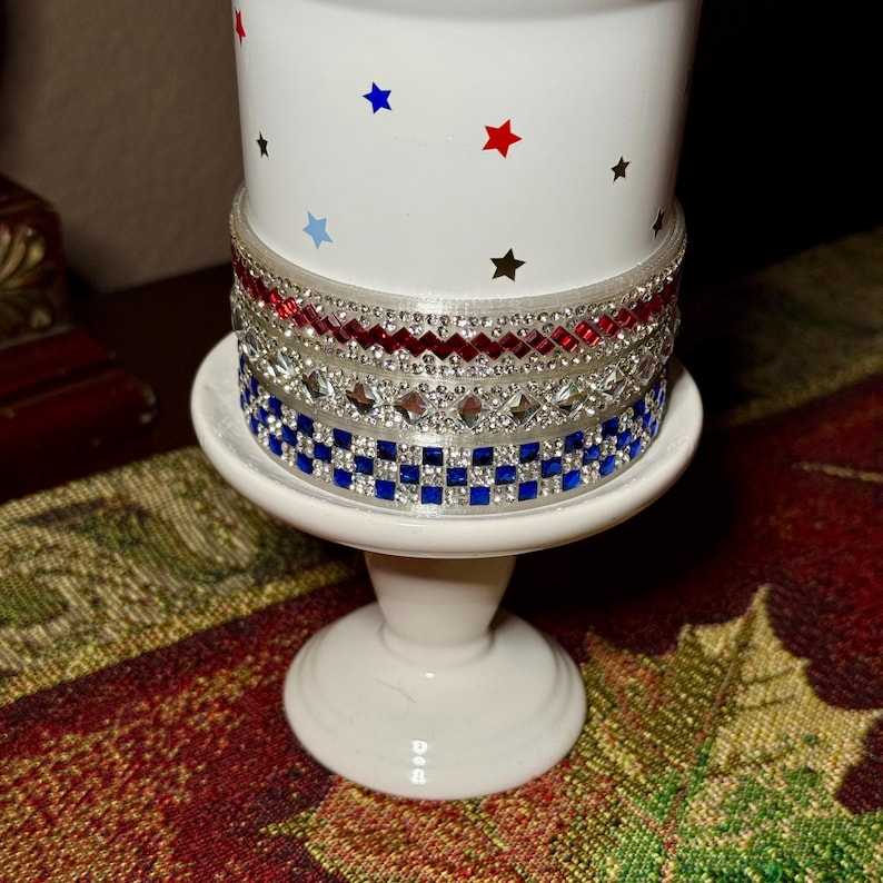 May include: A white candle holder with a decorative design. The candle holder has a white base and a cylindrical body adorned with red, blue, and silver embellishments. The top of the candle holder has red, blue, and black star accents.