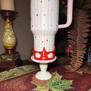 May include: A white tumbler with a pink handle, decorated with red, white, and blue stars and stripes. A red star-shaped crown sits on a white pedestal, adding a patriotic touch. The tumbler is approximately 7 inches tall.