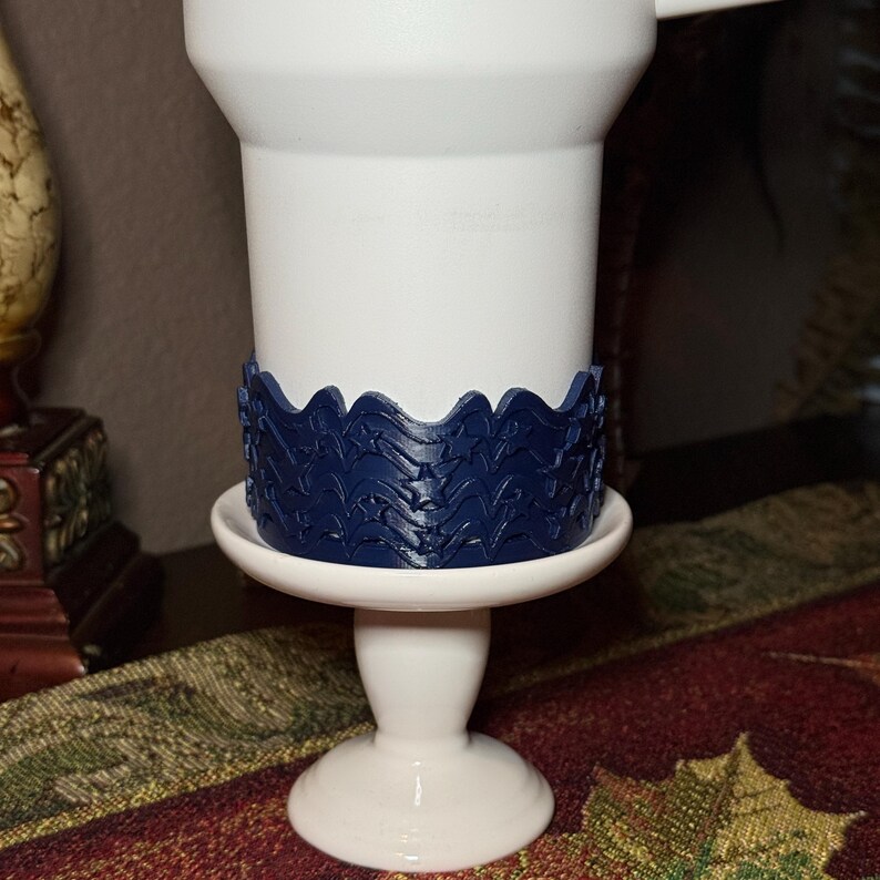 May include: A white pedestal cup featuring a dark blue, textured band wrapped around its middle section. The band displays a repeating pattern of stars and wave-like lines. The cup has a smooth, matte finish and stands on a circular base.