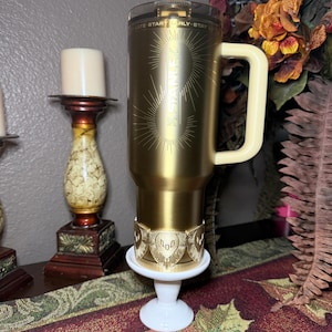 May include: A gold-colored travel mug with a cream-colored handle and a clear lid. The mug has the text "STAINER" and a sunburst design. It sits on a white pedestal with a gold-colored decorative band.