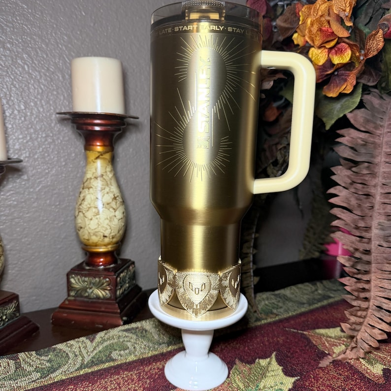 May include: A gold-colored travel mug with a cream-colored handle and a clear lid. The mug has the text "STAINER" and a sunburst design. It sits on a white pedestal with a gold-colored decorative band.