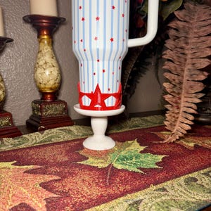 May include: A white tumbler with blue stripes and red stars, paired with a red crown-shaped coaster. The tumbler sits on a white pedestal, with a decorative table runner in the foreground. A candle holder is visible in the background.