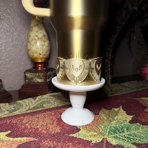 May include: A gold-colored tumbler with a decorative band featuring a shield design. The tumbler sits on a small, white, pedestal-style stand. The background includes a decorative vase and a patterned tablecloth.