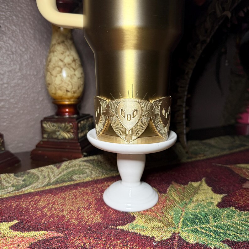 May include: A gold-colored tumbler with a decorative band featuring a shield design. The tumbler sits on a small, white, pedestal-style stand. The background includes a decorative vase and a patterned tablecloth.