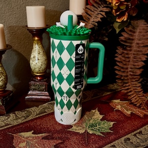 May include: A white and green insulated tumbler with a golf theme. The tumbler features a green diamond pattern, golf ball and tee graphics, and a green handle. The lid has a green grass design with a golf ball and tee. The tumbler is designed to keep drinks cold for up to 24 hours.