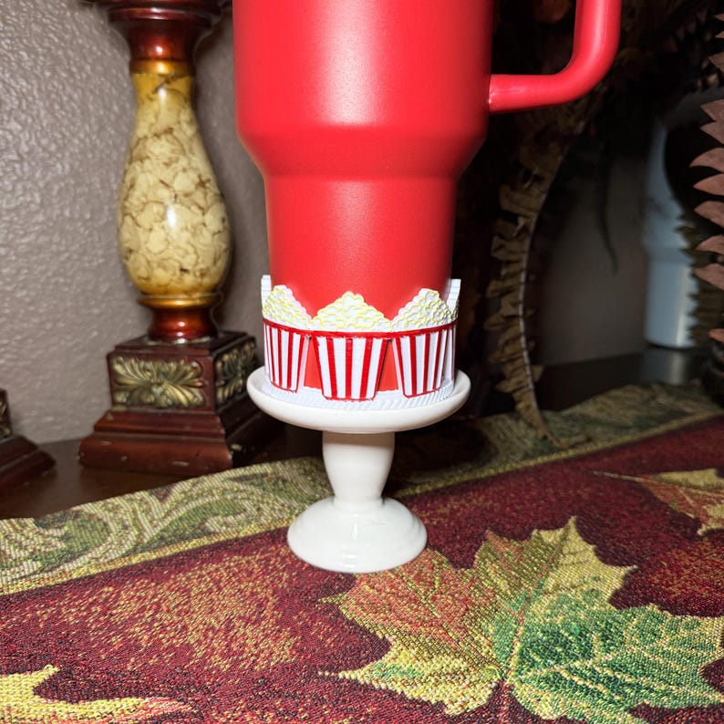 Pu&ograve; includere: Un bicchiere rosso brillante con una fascia in silicone avvolgente decorata con un motivo ripetitivo di secchielli per popcorn a righe rosse e bianche pieni di popcorn gialli, poggiato su un piccolo supporto bianco con base circolare.