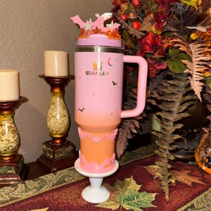 May include: A pink and orange Stanley tumbler with a handle, decorated with bats and stars. The tumbler sits on a white pedestal. The Stanley logo is visible. The lid is decorated with bats and stars.
