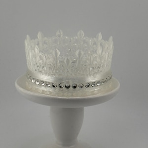May include: A white cake topper shaped like a crown with a clear, sparkly finish. The crown is decorated with a row of silver rhinestones around the base.