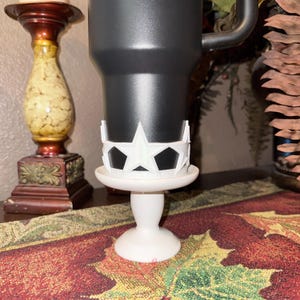 May include: A black tumbler sits on a white pedestal with a star-shaped design. The pedestal has a round base and a slender stem. The star design is white and surrounds the tumbler. The background includes a decorative candle holder and a fall-themed table runner.