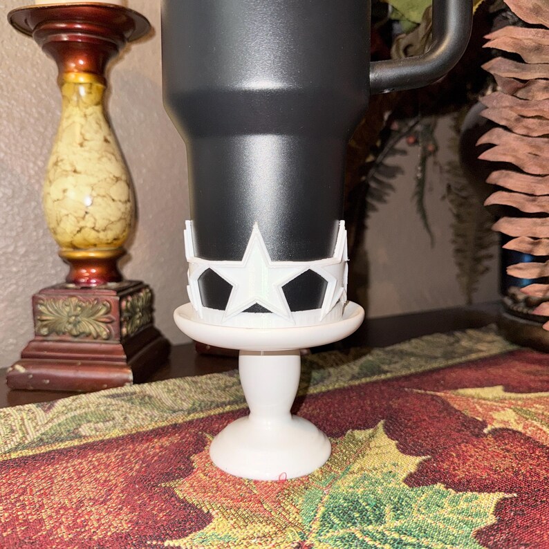 May include: A black tumbler sits on a white pedestal with a star-shaped design. The pedestal has a round base and a slender stem. The star design is white and surrounds the tumbler. The background includes a decorative candle holder and a fall-themed table runner.
