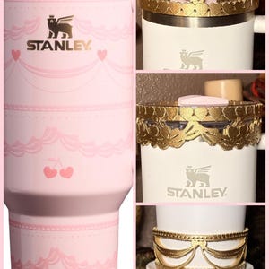May include: Pink Stanley tumbler with a decorative design featuring cherries and hearts. The tumbler has a straw and a lid. The Stanley logo is visible. The image also shows a white mug with gold decorative bands.
