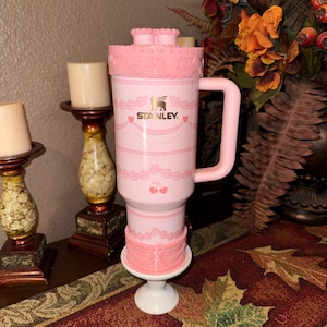 May include: A pink Stanley travel mug with a cake-themed design. The mug has a handle, a decorative lid, and a base. The Stanley logo is visible. The design includes cherries and decorative trim.