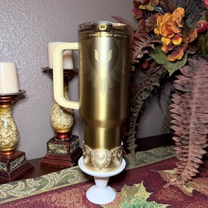 May include: A gold-colored travel mug with a cream-colored handle and lid. The mug features a sunburst design with a central emblem and the text "START EARLY - STAY LATE". It sits on a small white pedestal.