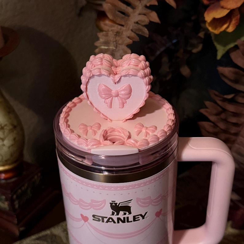 May include: Pink Stanley travel mug with a decorative heart-shaped lid. The lid features a bow design and a scalloped edge. The mug has a handle and the Stanley logo. The mug is decorated with hearts and bows.
