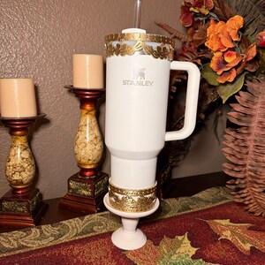 May include: A white Stanley tumbler with a gold leaf design around the top and base. The tumbler has a handle and a straw. Two candle holders with candles are on the left. A fall floral arrangement is in the background.