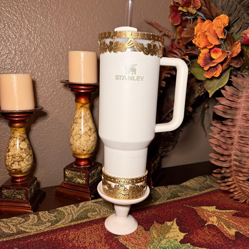 May include: A white Stanley tumbler with a gold leaf design around the top and base. The tumbler has a handle and a straw. Two candle holders with candles are on the left. A fall floral arrangement is in the background.