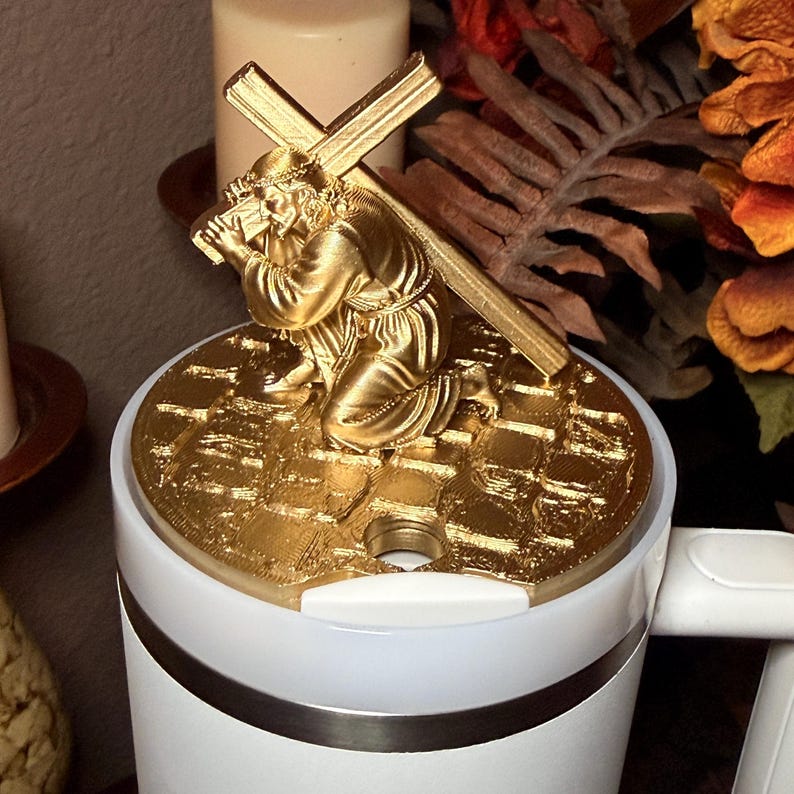 Puede incluir: Figura religiosa dorada que representa una figura arrodillada que lleva una cruz. La figura est&aacute; montada sobre una base blanca y plateada. El fondo incluye una vela y follaje oto&ntilde;al.