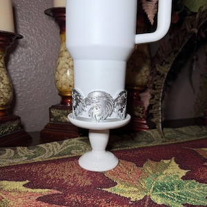 May include: A white travel mug with a silver eagle design around the base, set on a white pedestal. The mug has a handle and is surrounded by decorative elements, including a patterned runner and candle holders.