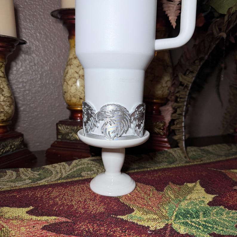 May include: A white travel mug with a silver eagle design around the base, set on a white pedestal. The mug has a handle and is surrounded by decorative elements, including a patterned runner and candle holders.
