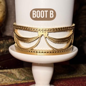 May include: A white ceramic candle holder with a gold-colored decorative band. The band features a detailed design of draped elements. The text "BOOT B" is displayed on the candle holder.