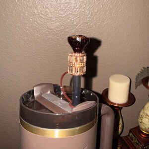 May include: A brown and gold travel mug with a decorative straw. The straw features a sparkling, faceted, dark brown gem at the top, with a gold and crystal band. The mug has a brown handle and a gold rim.