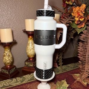 May include: White insulated water bottle with a black band featuring the word "COLDEST" and "LIMITLESS SERIES". The bottle has a white handle and a black textured top and bottom. A white straw extends from the top.