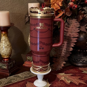 May include: A maroon Stanley travel mug with a gold-colored crown-like lid and base. The mug has a handle and is decorated with a lace-like pattern and cherry illustrations. The Stanley logo is visible on the front.