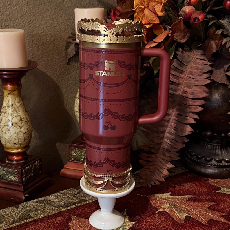 May include: A maroon Stanley travel mug with a gold-colored crown-like lid and base. The mug has a handle and is decorated with a lace-like pattern and cherry illustrations. The Stanley logo is visible on the front.