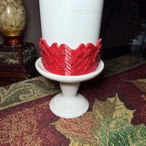 May include: A white pedestal stand featuring a cylindrical base and a wide, flat top. A bright red, textured band with a repeating pattern of stars and wavy lines is wrapped around the lower portion of the white cylinder resting on the stand.