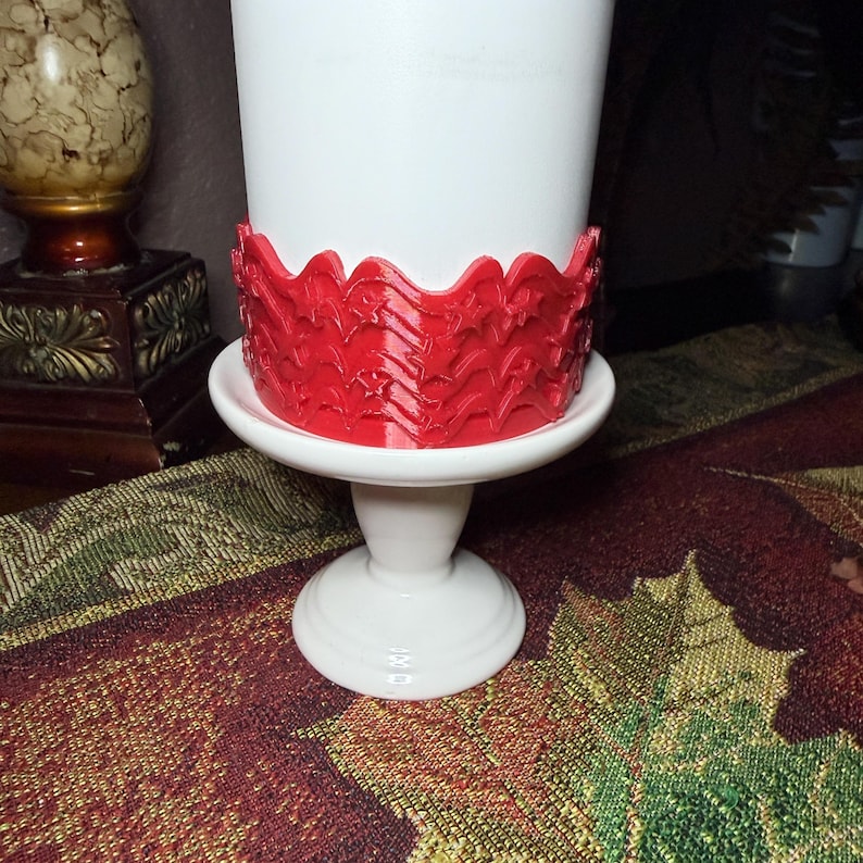 May include: A white pedestal stand featuring a cylindrical base and a wide, flat top. A bright red, textured band with a repeating pattern of stars and wavy lines is wrapped around the lower portion of the white cylinder resting on the stand.