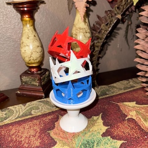 May include: A stack of three-dimensional star-shaped crowns in red, white, and blue, resembling the American flag. The crowns are stacked on a small, white pedestal. The crowns are made of plastic.