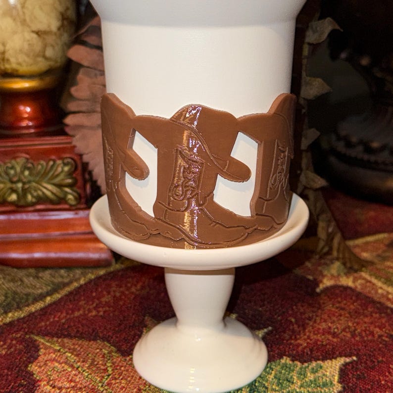 K&ouml;nnte beinhalten: Ein wei&szlig;er Eierbecher aus Keramik mit einem braunen Cowboy-Stiefel-Design am oberen Rand. Der Eierbecher steht auf einem wei&szlig;en Sockel. Das Design ist ein sich wiederholendes Muster von Cowboy-Stiefeln.