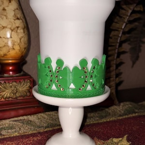May include: White candle holder with a green decorative band featuring shamrocks and horseshoes, adorned with small gold-colored accents. The candle holder sits on a white pedestal base, creating a festive and decorative item.
