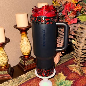 May include: A black Stanley travel mug with a red rose-shaped lid. The mug has a black handle and is decorated with red and gold accents. It is displayed on a white pedestal.