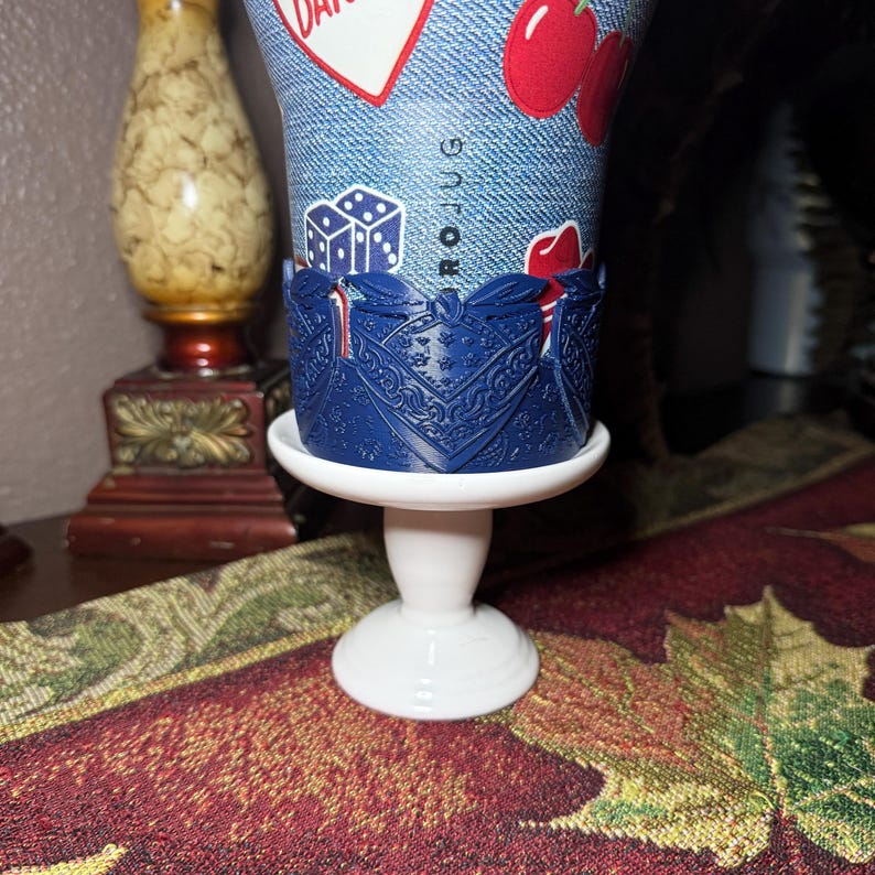 May include: A tumbler with a blue denim-textured surface featuring red cherry and white heart graphics, along with dice icons and the text ROJUG. The tumbler is held by a dark blue, intricate, laser-cut bandana-patterned sleeve on a white pedestal.