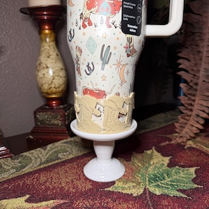 K&ouml;nnte beinhalten: Ein 1,14-Liter-Reisebecher aus Edelstahl mit wei&szlig;em Griff, der ein Western-Design mit Kakteen, Stiefeln und Langhorn-Rindern aufweist. Der Becher steht auf einem wei&szlig;en Sockel mit einem hellbraunen, stiefelf&ouml;rmigen Halter.