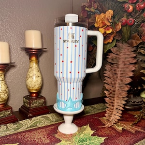 May include: A Stanley travel mug with a white handle, clear lid, and a blue and white striped pattern with red stars. The mug is on a white pedestal with a blue star-shaped base. The Stanley logo is visible.