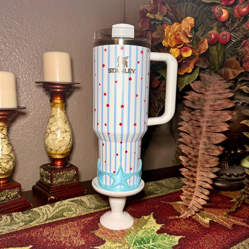 May include: A Stanley travel mug with a white handle, clear lid, and a blue and white striped pattern with red stars. The mug is on a white pedestal with a blue star-shaped base. The Stanley logo is visible.